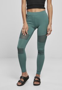 Leggings verdi con una superficie liscia, dotati di pannelli in rete nera alle ginocchia. Progettati per il massimo comfort con un elastico in vita alto.