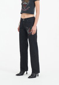 Black denim jeans with a straight leg design, featuring colorful embroidered details on the left thigh. Worn with a fitted graphic top and heeled shoes.