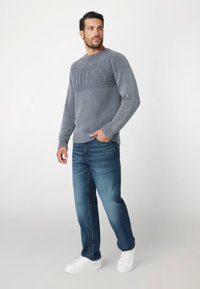 Maglione grigio a maglia con un design del logo in rilievo, abbinato a jeans in denim blu e scarpe da ginnastica bianche, creando un aspetto casual.