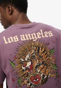 Camiseta de algodón morada con un estampado gráfico de un tigre rugiente, rodeado de hojas y un corazón, con el texto "los angeles" encima.