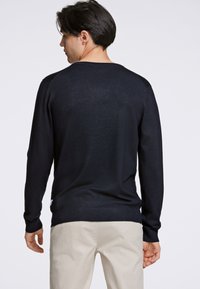 Maglione blu navy con maniche lunghe, orlo e polsini a costine, texture liscia, design aderente, indossato sopra pantaloni chiari.