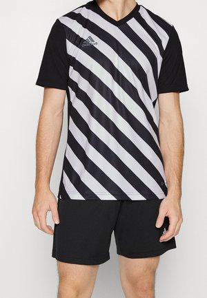 Mann trägt ein schwarz-weiß diagonal gestreiftes Adidas-Sportshirt mit schwarzen Ärmeln und schwarzen Shorts vor einfarbigem Hintergrund.