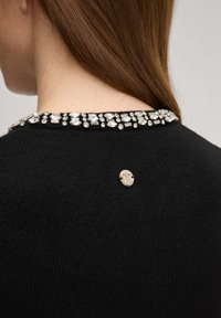 Μαύρο πουλόβερ με λαμπερή κρυστάλλινη διακόσμηση στο ντεκολτέ και ασημένια λεπτομέρεια υλικού στο πίσω μέρος. Ύφασμα πλεκτό με υφή.