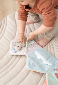 Peuter in oranje shirt knielt op een beige mat en tilt een stoffen klep op van een zacht interactief speelgoedboek met een pluchen dier erin.