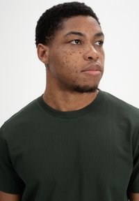 Donkergroene katoenen t-shirt met een ronde hals en korte mouwen. Gladde textuur zonder zichtbare patronen of grafische elementen. Eenvoudig ontwerp.