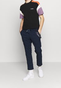 Camiseta negra con mangas multicolores (naranja, crema, púrpura) combinada con pantalones de cuadros azul marino y zapatillas blancas con superficie texturizada.