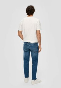 Moška belo majica s kratkimi rokavi v kombinaciji z modrimi jeans hlačami, ki imajo zadnje žepe in teksturirano tkanino, oblečena z belimi športnimi copati.