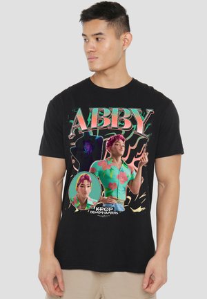Young man wearing black tee with colorful "ABBY" and animated character images from KPOP Demon Hunters, standing against plain background.