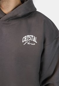 Felpa grigia con cappuccio, dotata di tasca frontale, polsini a coste e un logo ricamato bianco con la scritta "CRYSTAL Paris" sul petto.