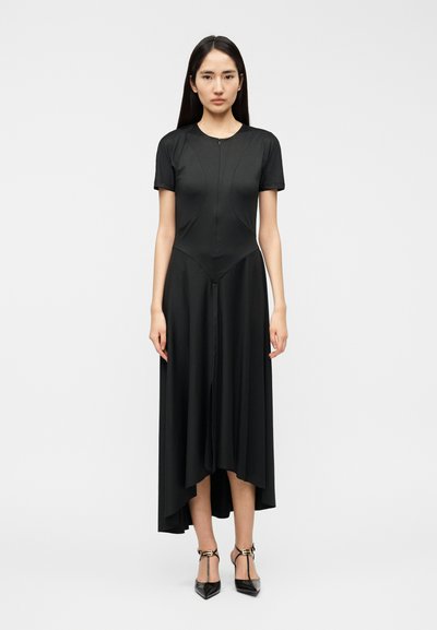 Vestido negro de manga corta con corpiño ajustado, silueta de línea A, dobladillo asimétrico y textura suave, combinado con tacones negros puntiagudos.
