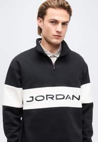 Νέος άνδρας φορώντας μαύρο και άσπρο φούτερ με φερμουάρ ως το στήθος και τη λέξη «JORDAN» στο στήθος, στέκεται μπροστά σε απλό φόντο.