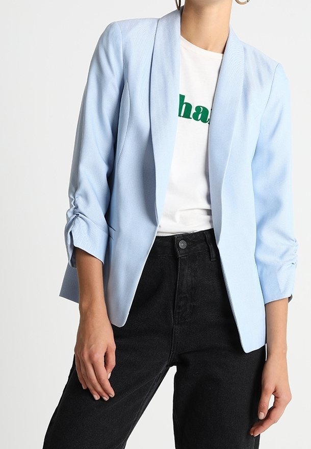 Femme portant un blazer bleu clair sur un t-shirt blanc à motif graphique et un jean noir taille haute, debout devant un fond uni.