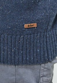 Pull en laine bleu marine avec un motif texturé agrémenté de touches de bleu clair. Une étiquette en cuir brun avec le logo "Le Cooper" en bas.