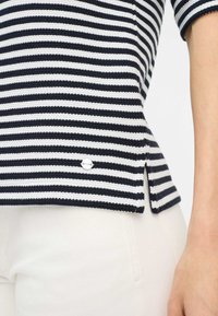 Gestreepte gebreide top met marineblauwe en witte horizontale strepen, met een zijsplit en een klein merkje aan de zoom. Gecombineerd met witte jeans.
