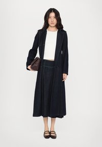 Mørk denimkåpe med grønn søm, kombinert med en tettsittende hvit topp og et langt denimskjørt. Tilbehør med en brun skulderveske og svarte sko.