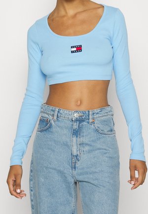 Nainen pukeutuneena vaaleansiniseen pitkähihainen crop-topiin, jossa on "Tommy Jeans" -logo, ja korkeavyötäröisiin vaaleansinisiin farkkuihin, paljastaen vatsan.