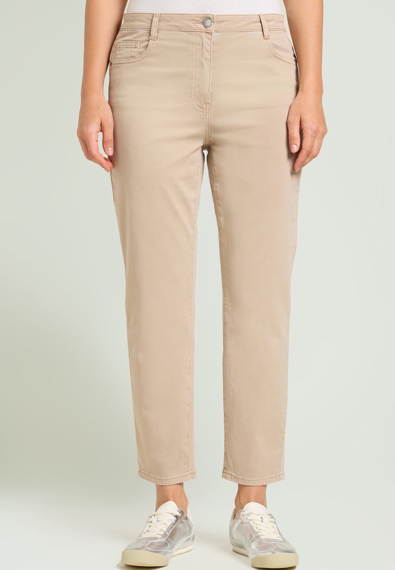 Pantalones beige a la altura del tobillo con corte recto, dos bolsillos frontales y cierre de botón. Combinados con zapatillas en tono plateado.
