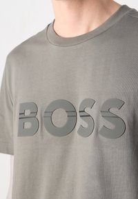 Homme portant un t-shirt gris clair avec de grandes lettres en relief formant "BOSS" sur la poitrine.