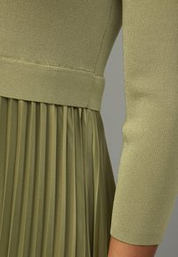 Abito verde oliva con un top in maglia a coste aderente e una gonna plissettata. La texture è liscia con una leggera lucentezza e un dettaglio in vita elastico.