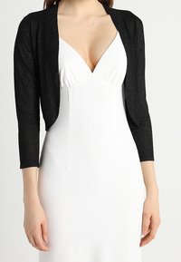 Cárdigan negro, corto, con mangas tres cuartos, de tejido texturizado, combinado con un vestido blanco de escote en V profundo. Sin hardware o patrones notables.
