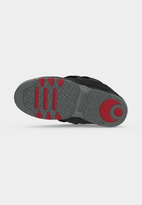 Semelle d'une chaussure de sport noire avec une bande de roulement grise et des accents rouges, présentant un motif texturé et un dessin circulaire rouge près du talon.