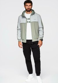 Lys grå og olivengrøn jakke med hætte, lynlås foran, elastiske manchetter; parret med sorte cargo-bukser og hvide sneakers, der har en t-shirt med logo-print.