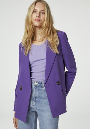 Paarse blazer met een dubbelbreasted ontwerp, gelapte revers, knopen en een gladde textuur, gecombineerd met een lichtpaarse geribde top en jeans.