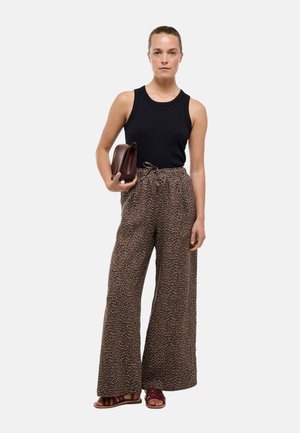 Femme portant un haut noir sans manches, un pantalon à jambes larges et à motif, des sandales marron, tenant une pochette marron, debout devant un fond uni.