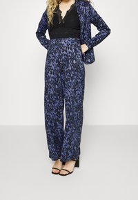 Jumpsuit leopard print blu navy con pantaloni larghi e top in pizzo nero. Presenta una vita aderente e maniche lunghe; abbinato a sandali neri con cinturini.