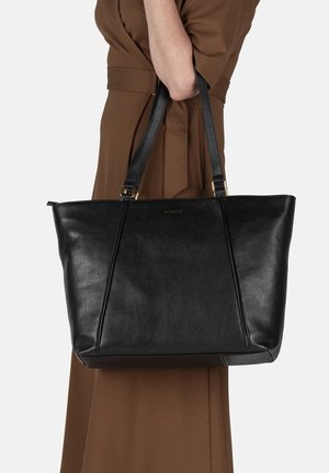 Donna che indossa un vestito marrone tiene una grande borsa tote in pelle nera con doppie spalline e un logo discreto sul davanti.