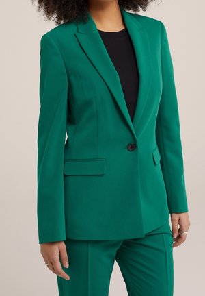 Blazer vert avec une coupe ajustée, fermeture à un bouton, revers crantés, poches latérales et texture de tissu lisse. Associé à un pantalon assorti.