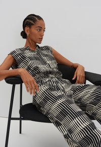 Black and cream jumpsuit with vertical and horizontal stripes, featuring a collar and sleeveless design. Worn while seated on a black chair.