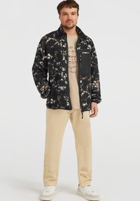 Fleecejacke in Schwarz mit weißen und beigefarbenen abstrakten Mustern, durchgehender Reißverschluss, hoher Kragen und Brusttasche. Getragen über einem beige T-Shirt und khakifarbenen Hosen.