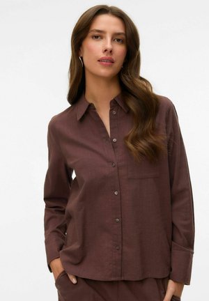 Hosszú, hullámos hajú nő laza barna gombos ingben és hozzáillő nadrágban, zsebeibe tett kézzel áll fehér háttér előtt.