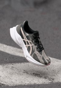 Laufschuh mit einem Obermaterial aus schwarzem und beigem Mesh mit gestreifter Textur, schlanker weißer Zwischensohle und schwarzen Schnürsenkeln, der über einer Fläche schwebt.