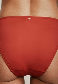Červené spodní díly bikini vyrobené z hladké tkaniny. Vyznačují se stříbrným kovovým akcentem na zadní straně s čistým, minimalistickým designem a jednoduchým šitím.