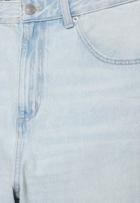 Zbliżenie na jasnoniebieskie jeansy z denimu pokazujące przednią kieszeń, szlufkę na pasek, zamek błyskawiczny oraz metalowy guzik z grawerem marki.