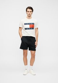 Λευκό βαμβακερό μπλουζάκι με μπλε και κόκκινο γραφικό λογότυπο "TOMMY JEANS", συνδυασμένο με μαύρο σορτς και λευκά αθλητικά παπούτσια. Απλό φόντο.