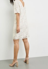 Vestido blanco con un patrón de crochet texturizado, escote en V y mangas cortas. Combinado con sandalias de tacón alto en color beige.