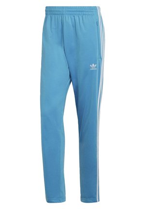 Mann trägt hellblauen Adidas-Trainingsanzug mit weißen Streifen und weißen Turnschuhen, steht vor einfarbigem Hintergrund.