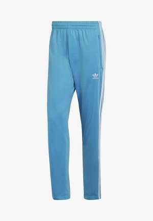 Mann trägt hellblauen Adidas-Trainingsanzug mit weißen Streifen und weißen Turnschuhen, steht vor einfarbigem Hintergrund.