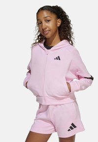 Sudadera con cremallera de color rosa claro con acentos y logotipo negros, combinada con pantalones cortos rosa a juego. Tejido suave, corte casual y diseño con capucha.