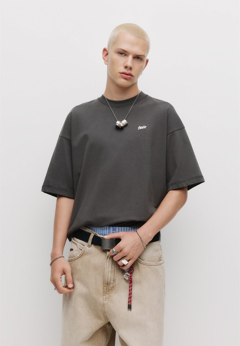 Mørkegrå oversized t-shirt med korte ærmer, rund hals og lille logo. Bæret med lysebeige jeans og et sort bælte, samt tilbehør.