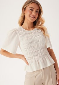 Blusa bianca in tessuto strutturato con maniche a palloncino corte, dettagli arricciati e orlo svasato. Tessuto liscio e leggera lucentezza.