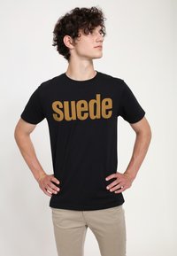 Černé tričko s velkým tučným nápisem "suede" v zlaté barvě. Krátké rukávy, kulatý výstřih, měkká látka. Nosené s světlými kalhotami.