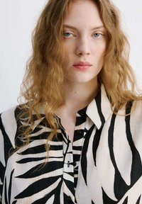 Jeune femme aux longs cheveux roux ondulés, portant une chemise boutonnée rayée zèbre noire et blanche, faisant face avec une expression neutre.