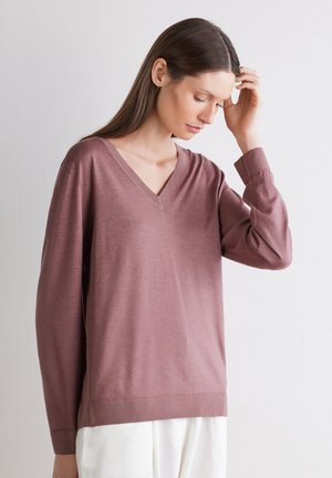 Kvinde med langt brunt hår iført en lilla V-hals sweater og hvide bukser, rører ved hendes hår mod en neutral grå baggrund.