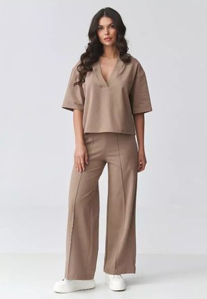 Kobieta o długich ciemnych włosach ubrana w luźną beżową bluzkę z dekoltem w V i pasujące szerokie spodnie, stojąca na tle jasnej, jednolitej ściany, w białych butach.