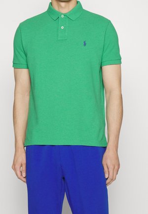Homme portant un polo vert à manches courtes avec deux boutons et un petit logo brodé, associé à un pantalon bleu sur un fond uni.