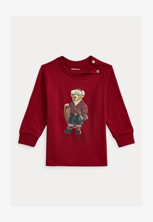 Camisola vermelha de manga comprida com uma estampa de urso vestido com roupas de inverno, detalhes em padrão xadrez e um trenó, com fecho de pressão no ombro.